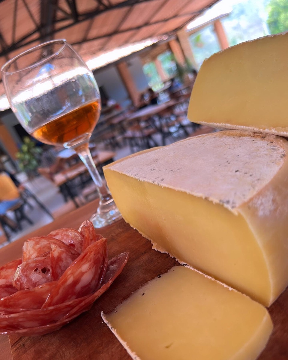 Queijo, salame fatiado e uma taça sobre tábua de madeira, com o salão do restaurante ao fundo